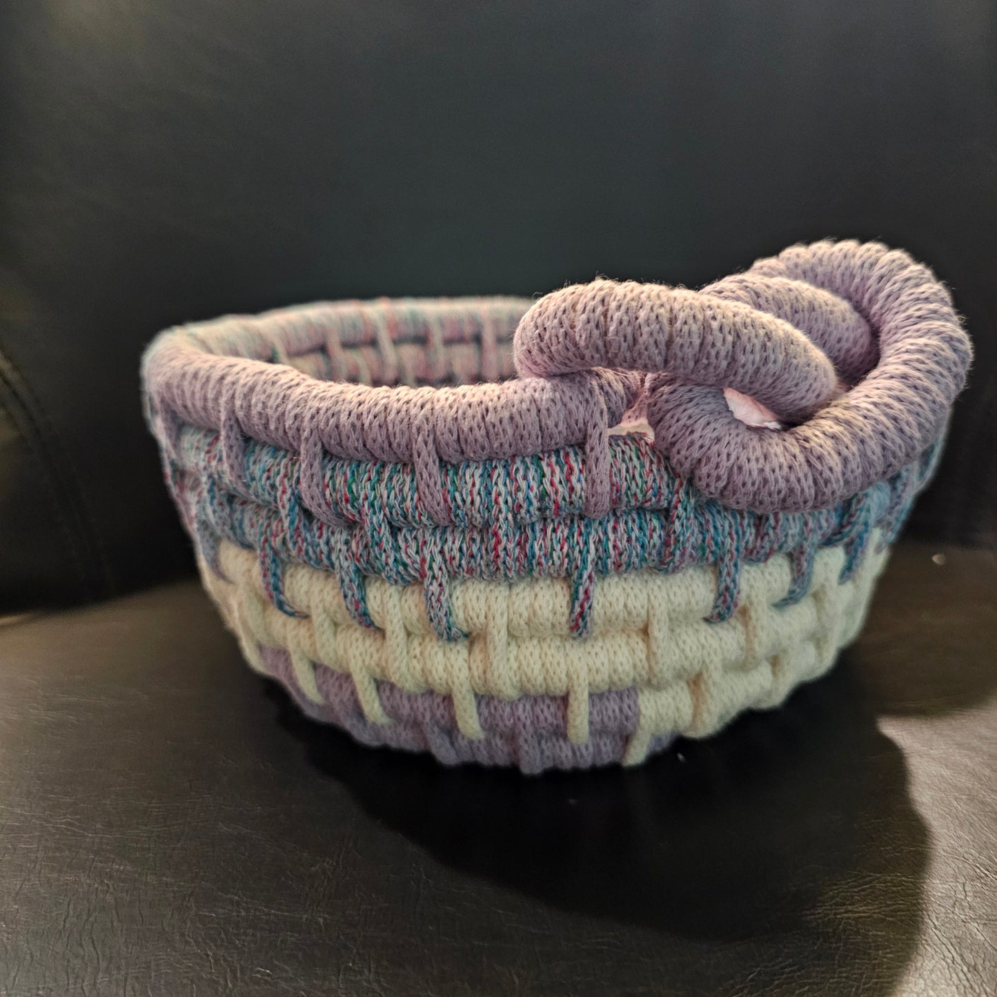 A small handwoven basket with a twisted loop handle, made from variegated yarn in lavender, cream, and light blue, resting on a dark leather surface.