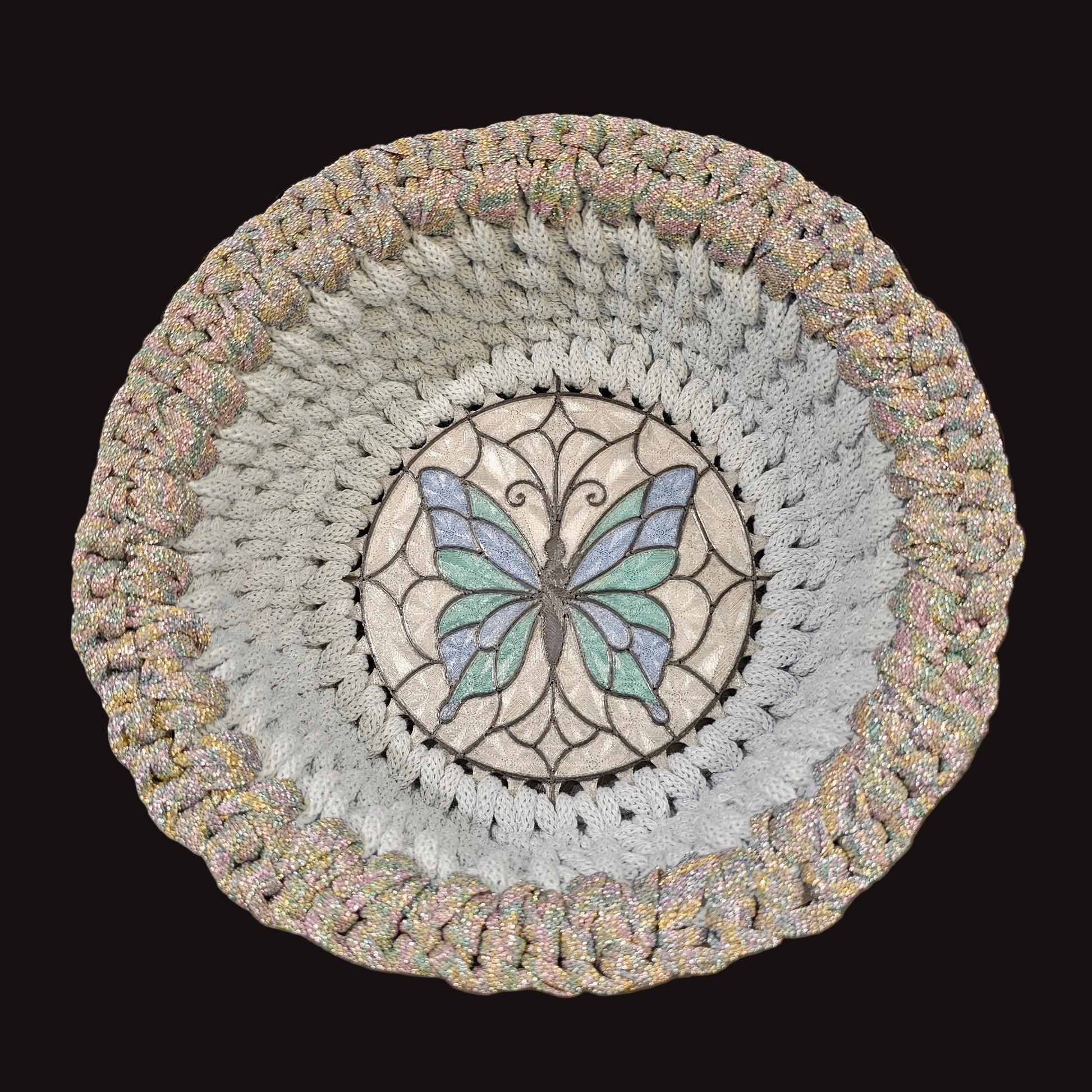 A round crocheted basket featuring a central stained-glass style butterfly motif in light blue and green, surrounded by concentric rings of light blue and a textured iridescent border. 9x9x3.5 inches overall.