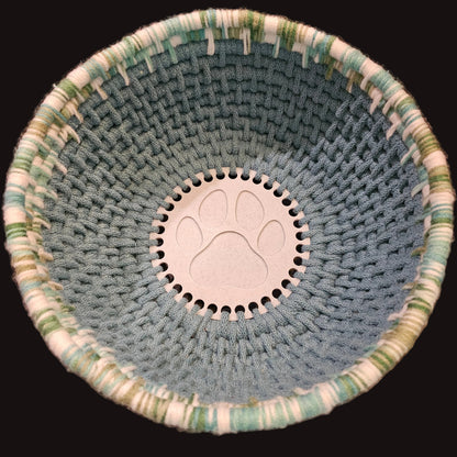A handmade coiled basket made from blue cord and varigated green and white yarn, featuring a white circular center with an embossed paw print.