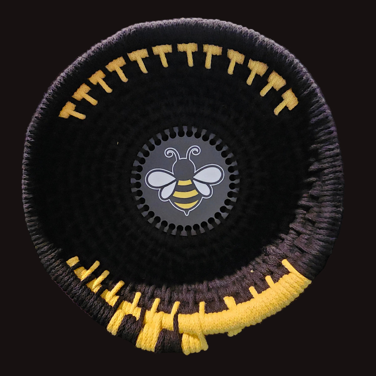 A black woven basket with yellow and black striped accents, featuring a circular black center insert with a white and yellow bee logo