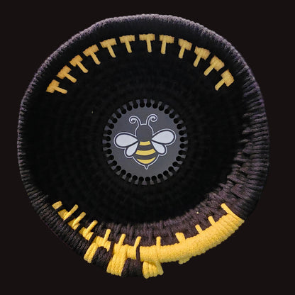 A black woven basket with yellow and black striped accents, featuring a circular black center insert with a white and yellow bee logo