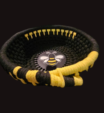 A black woven basket with yellow and black striped accents, featuring a circular black center insert with a white and yellow bee logo, set against a dark background.