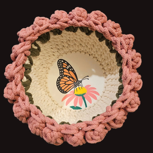 A round crocheted basket with a mauve tulip border, a cream center, and a butterfly and flower design at the base.