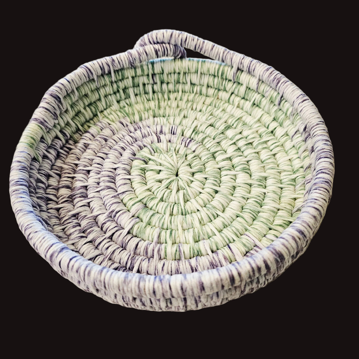 A round coiled basket with an integrated handle, made from striped white, light green, and purple fibers.