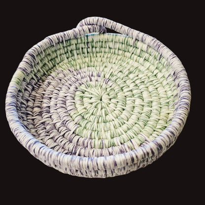 A round coiled basket with an integrated handle, made from striped white, light green, and purple fibers.