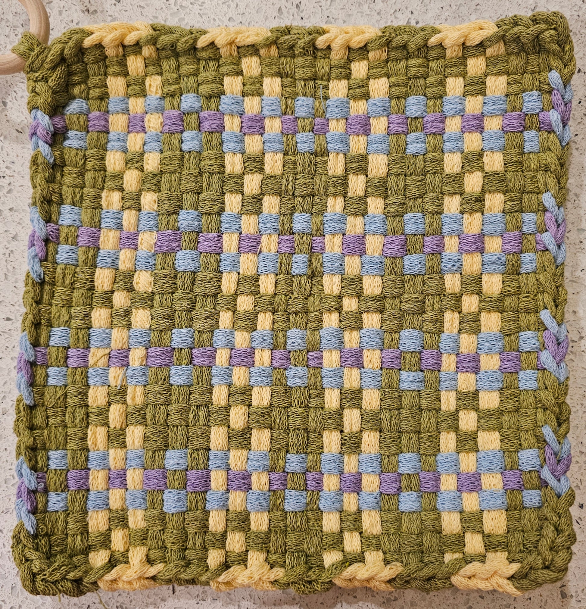 Woven mat with a checkered pattern in green, yellow, and purple on a textured surface.