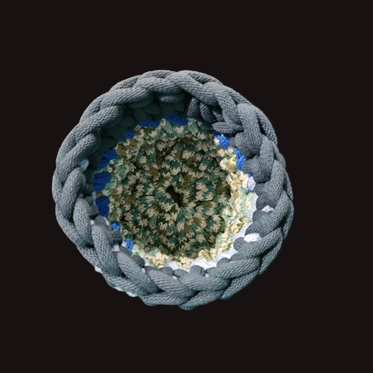 A handmade crocheted storage basket with a thick blue-gray outer rim and a multi-colored interior of green, teal, and cream yarn. Perfect for putting a potted plant in to add a touch of whimsy. 6x6x7.5 inches overall