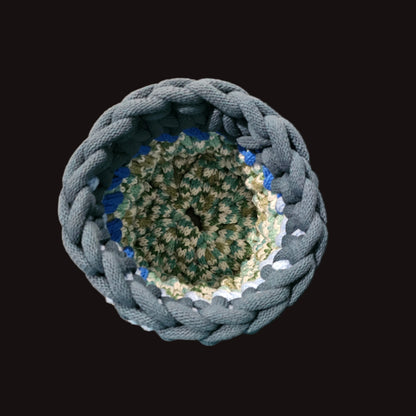 A handmade crocheted storage basket with a thick blue-gray outer rim and a multi-colored interior of green, teal, and cream yarn. Perfect for putting a potted plant in to add a touch of whimsy. 6x6x7.5 inches overall