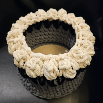 A handmade crocheted basket featuring a gray body, black leaves and a thick creamy braided cord featuring golden threads throughout forming tulips around the top. Approximately 8x8x6 inches overall