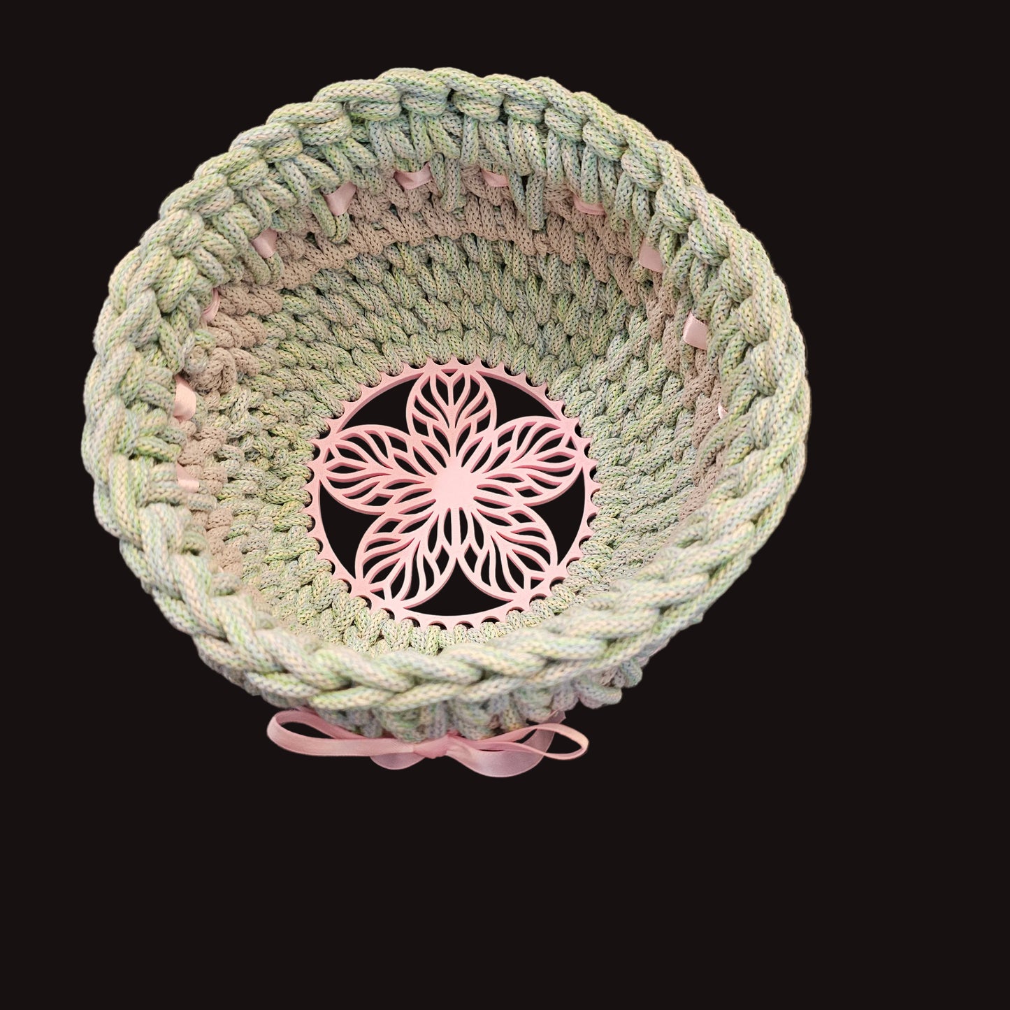 A handmade crocheted cord basket in pale green and pink, featuring a central pink floral cutout insert and a matching pink ribbon bow to finish it. 8x8x5 inches overall