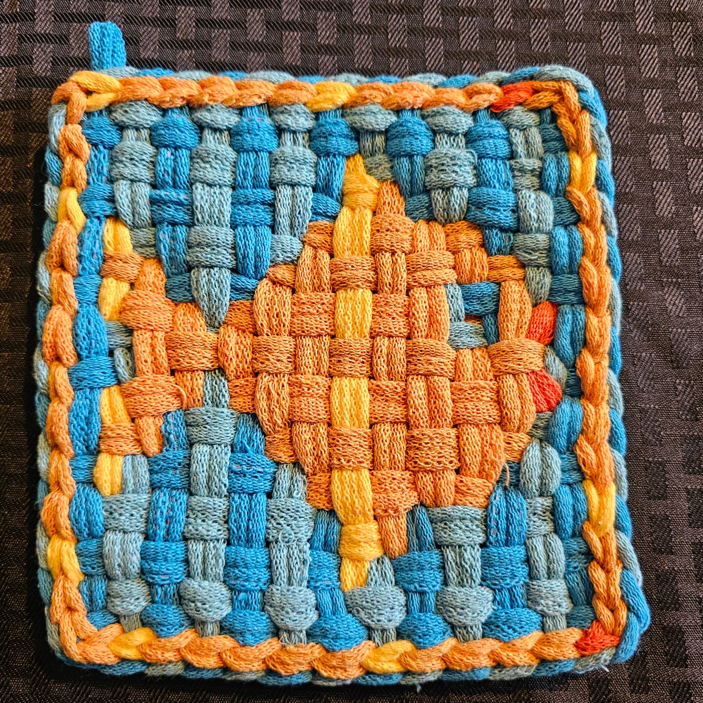 Woven mat with a fish design using blue, orange, and yellow yarns on a textured surface.