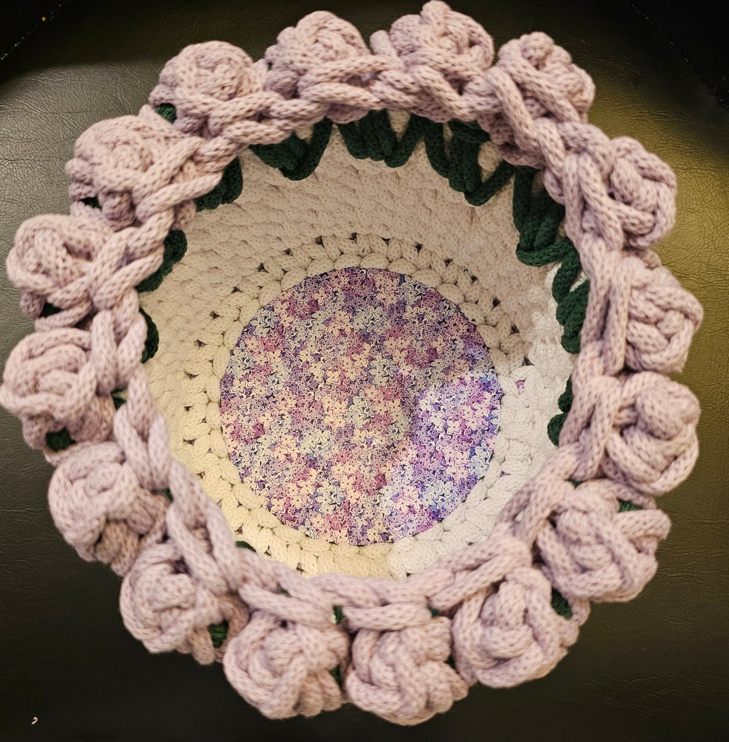 A crocheted basket in white with dark green leaves and soft lavender flowers around the top. At the center is a delicate but colorful purple and pink floral base. 7x7x5 inches overall