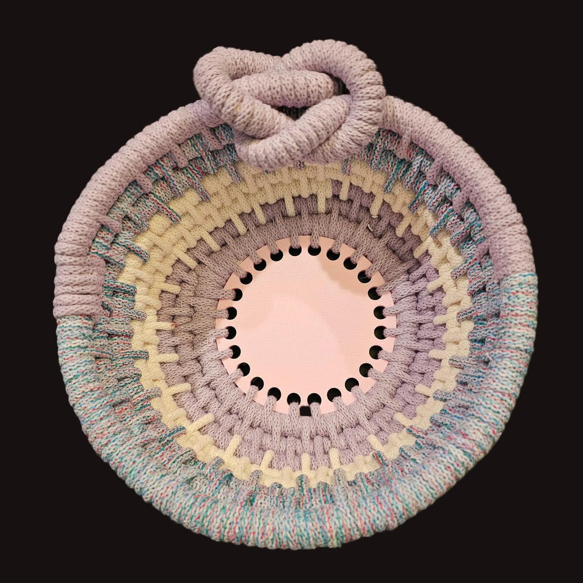 A round coiled basket with a thick knotted handle, featuring concentric rings of pastel lavender, cream, and variegated blue cord, photographed from above on a dark surface.