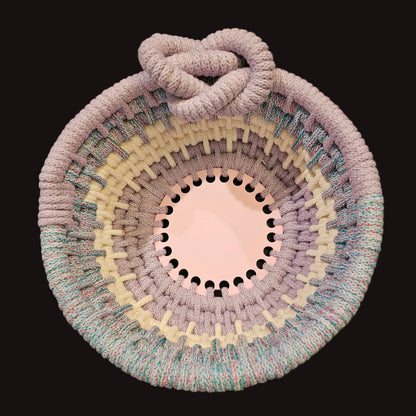 A round coiled basket with a thick knotted handle, featuring concentric rings of pastel lavender, cream, and variegated blue cord, photographed from above on a dark surface.