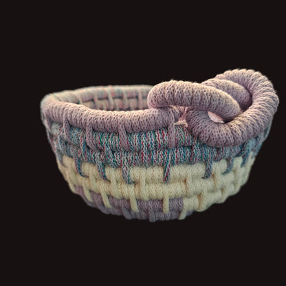 A small handwoven basket with a twisted loop handle, made from variegated yarn in lavender, cream, and light blue, resting on a dark leather surface.