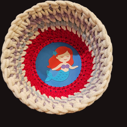 A handmade crocheted basket with concentric rings of sparkly cream, light blue, lavender, and red yarn, featuring a circular blue base with a sweet mermaid. 8x8x5 inches overall.