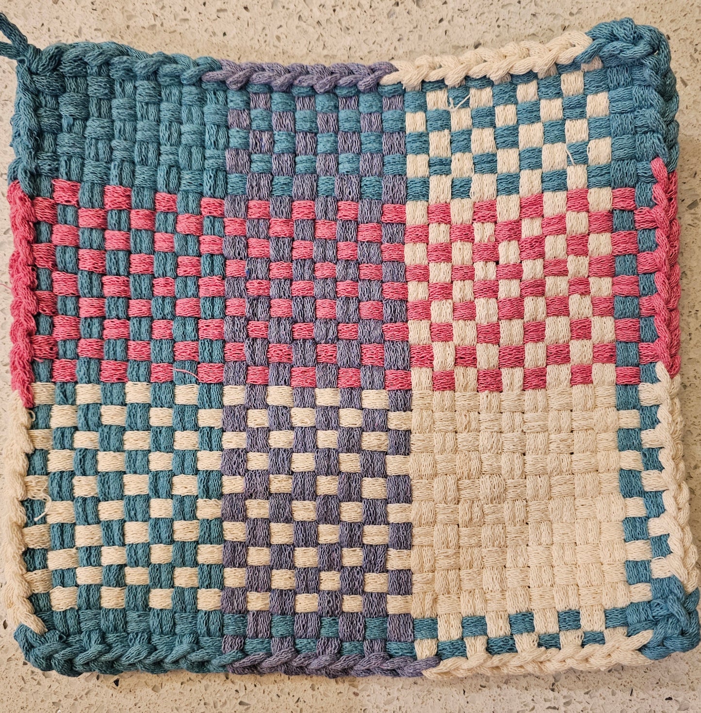 Woven mat with a checkered pattern in blue, pink, and white on a textured surface.