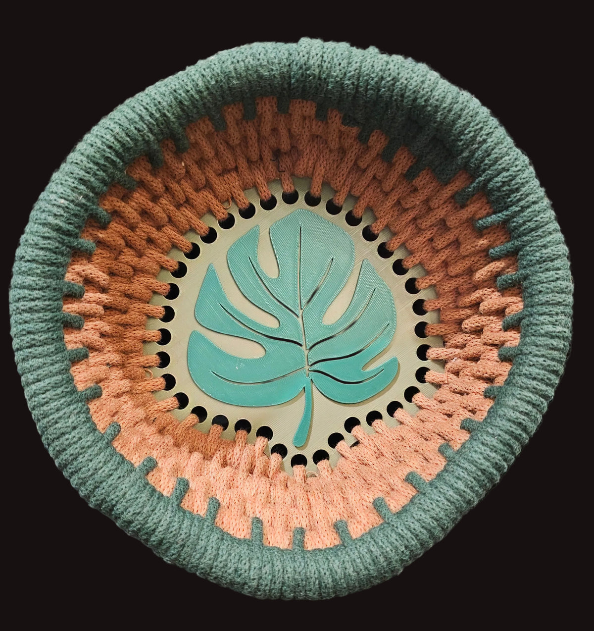 A round woven basket with a thick green braided rim and base, and a brown-colored woven body. The bottom center features a green monstera leaf design on a pale green background. 