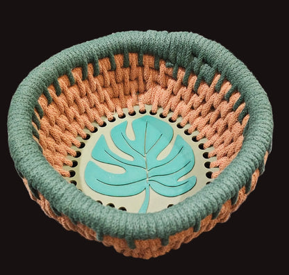 A round woven basket with a thick green braided rim and base, and a brown-colored woven body. The bottom center features a green monstera leaf design on a pale green background. 