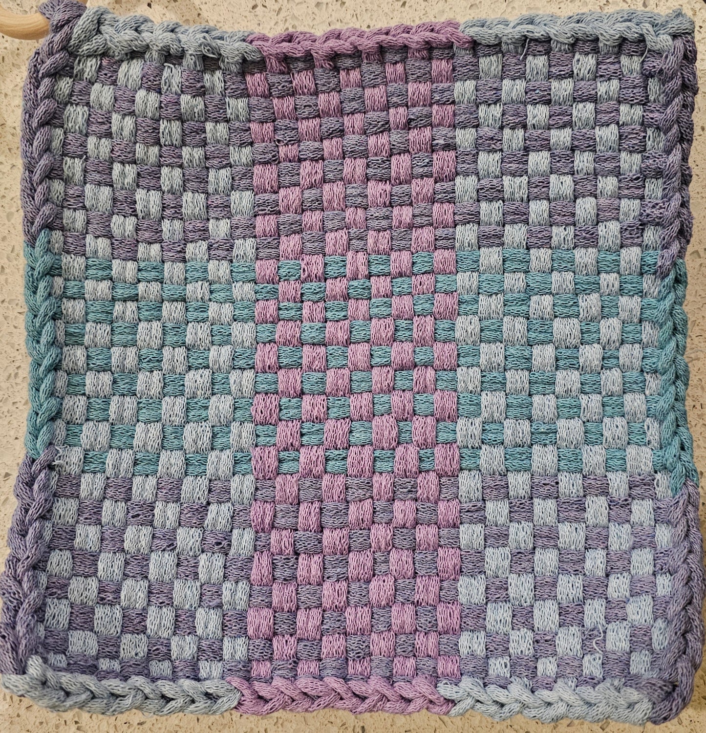 Woven fabric mat with a checkered pattern in purple, blue, and pink on a textured surface.