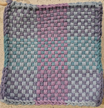 Woven fabric mat with a checkered pattern in purple, blue, and pink on a textured surface.