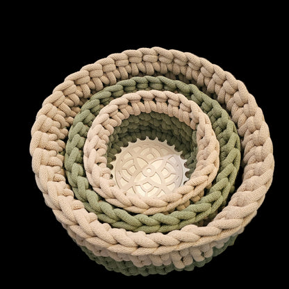 Three nested round woven baskets in natural beige, sage green, and olive green, with the largest basket resting on a black chair.