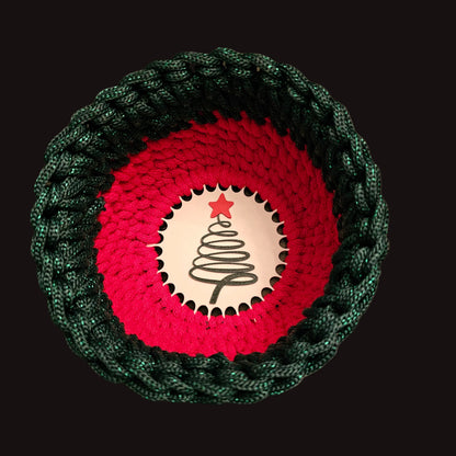A circular crocheted basket in Christmas red and finished off in a metallic sparkly deep green edge. At the center is a dark green spiral Christmas tree graphic with a red star.  6x6x4 inches overall