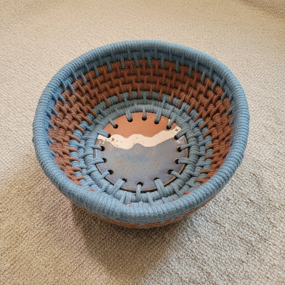 A circular, woven, rustic basket in concentric rings of steel blue and medium brown cord with a ceramic base.