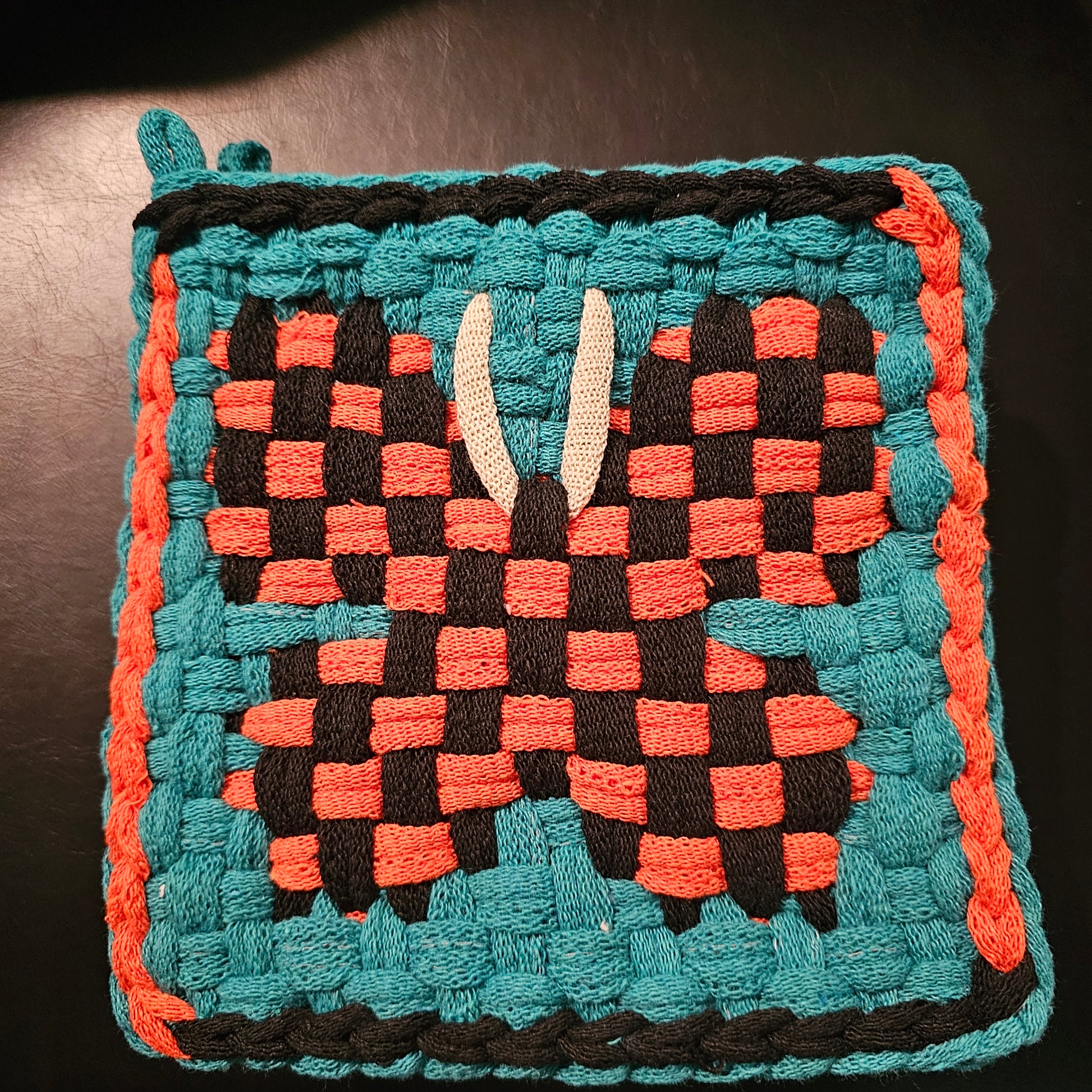 Woven square with a geometric pattern in teal, orange, and black on a dark background