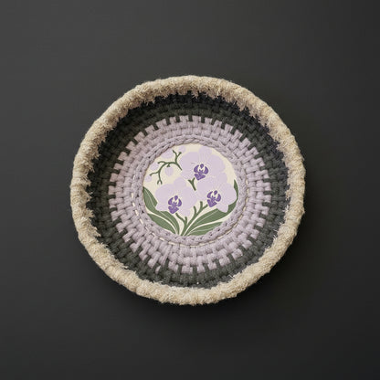 A round coiled basket with concentric rings of dark charcoal, gray, pale lavender, and natural beige frayed yarn, featuring a central floral medallion with purple orchids and green leaves on a cream background.
