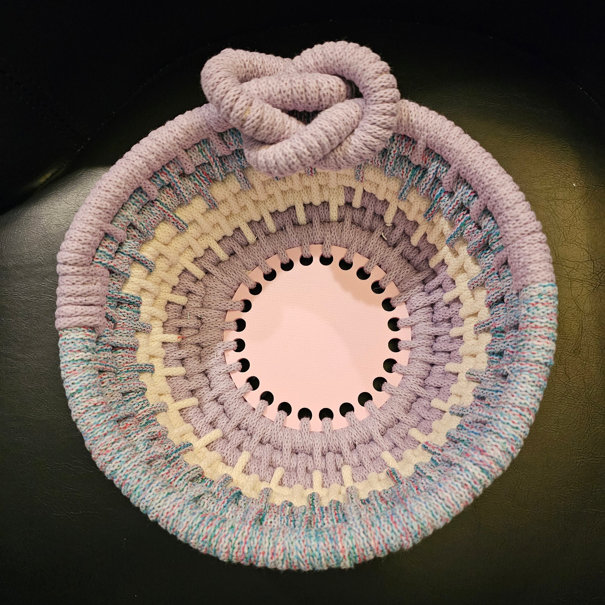 A round coiled basket with a thick knotted handle, featuring concentric rings of pastel lavender, cream, and variegated blue cord, photographed from above on a dark surface.