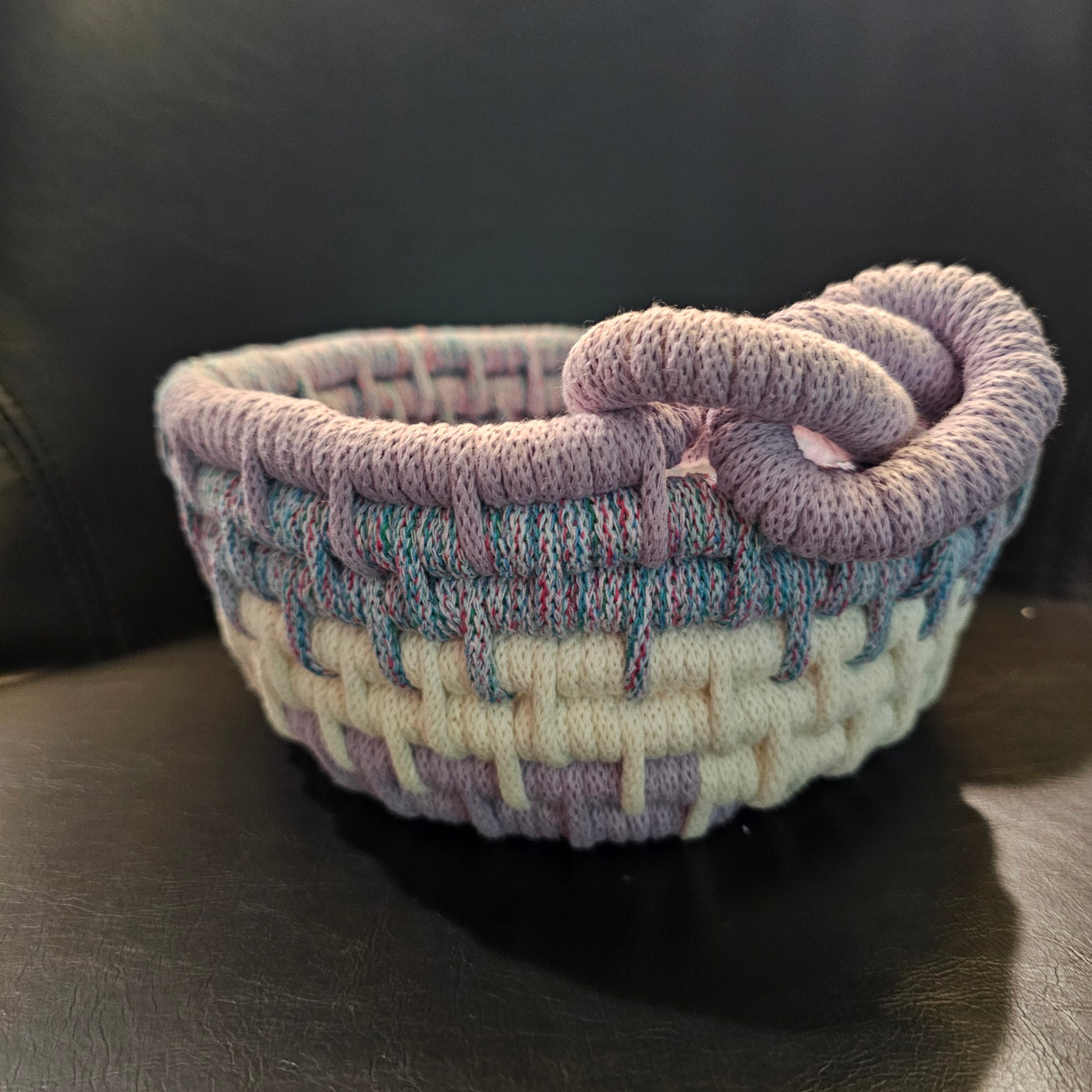 A small handwoven basket with a twisted loop handle, made from variegated yarn in lavender, cream, and light blue, resting on a dark leather surface.
