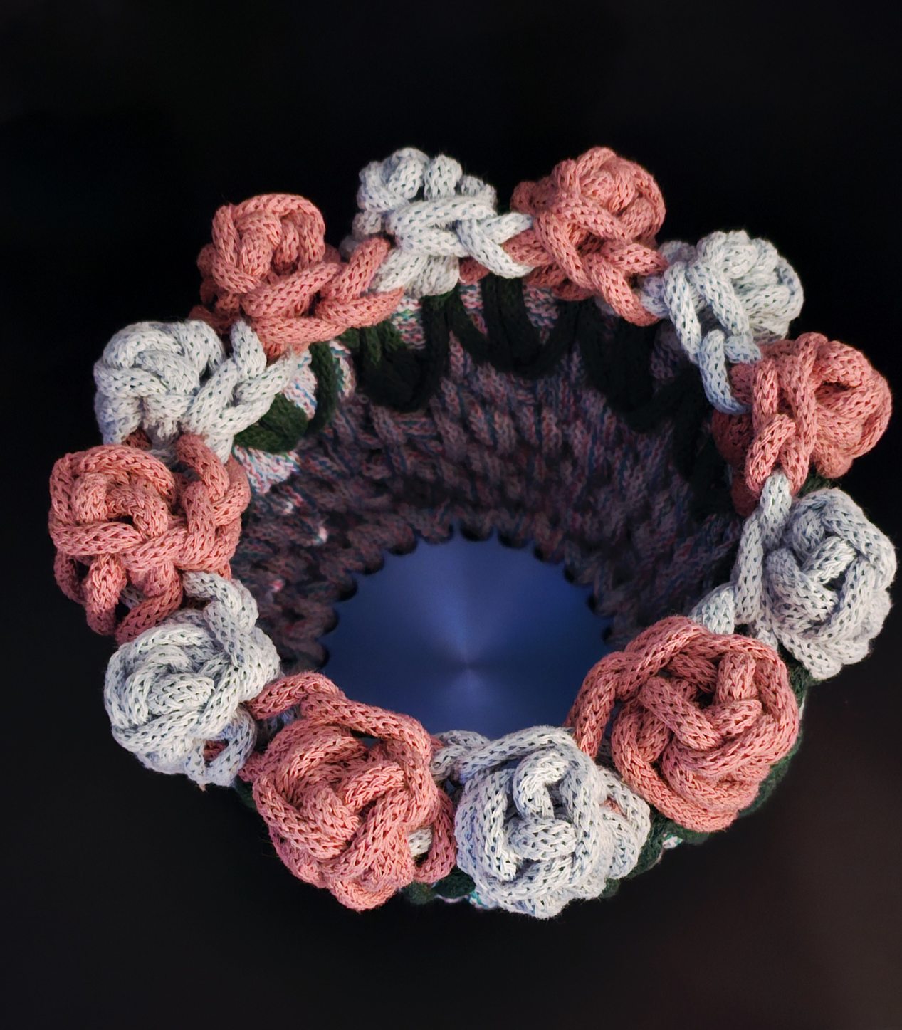 A handmade crocheted flower basket featuring alternating pink and light blue knotted roses with green leaves. It is all atop a variegated blue and pink cord attached to a bright shiny blue base. 7x7x5 inches overall.