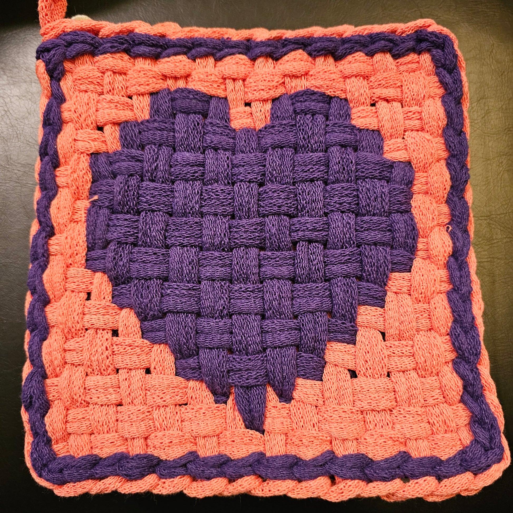 Hand-woven square mat with purple center and pink border on a dark background