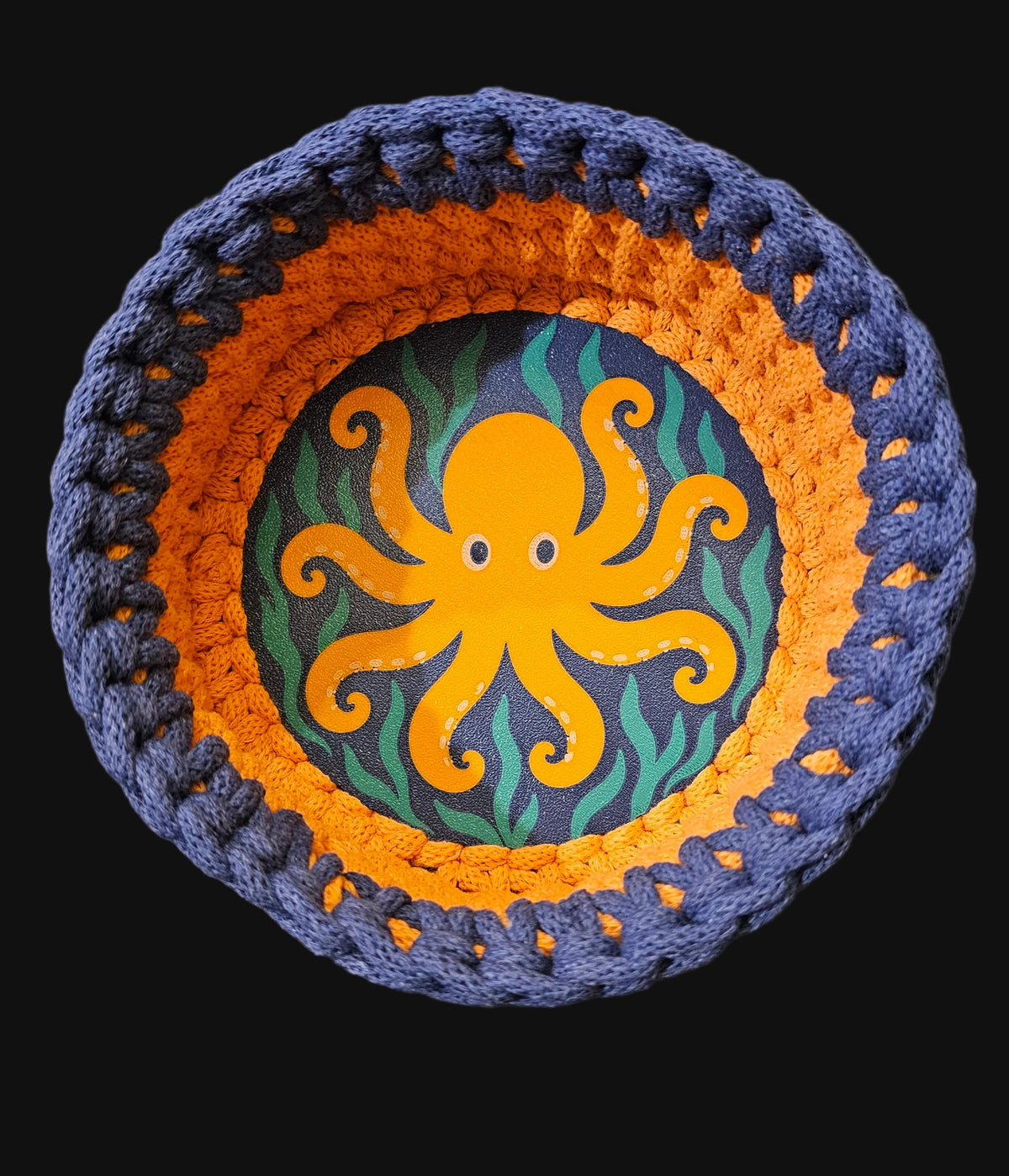 A round crocheted basket with an octopus design, featuring navy blue outer crochet trim, an orange middle band, and a dark blue center with a yellow-orange octopus and green seaweed pattern. 6x6x3 inches overall