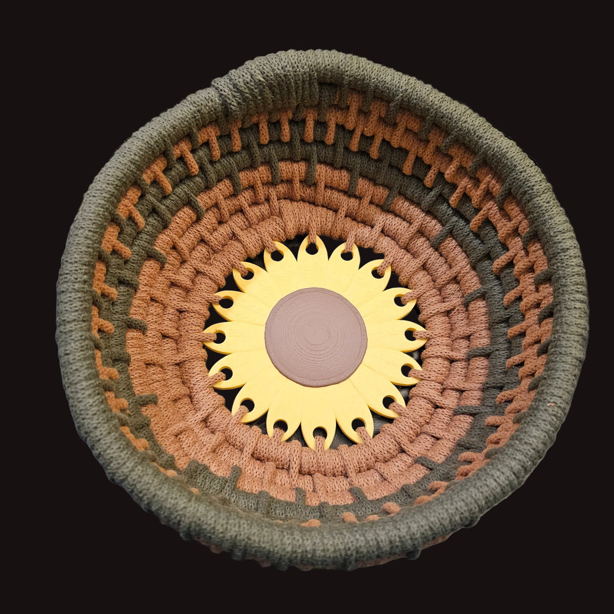 A handwoven basket with concentric rings of olive green, terracotta, and brown yarn, featuring a sunflower design in the center. 