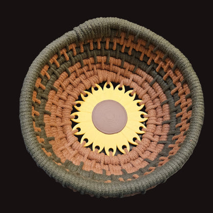 A handwoven basket with concentric rings of olive green, terracotta, and brown yarn, featuring a sunflower design in the center. 