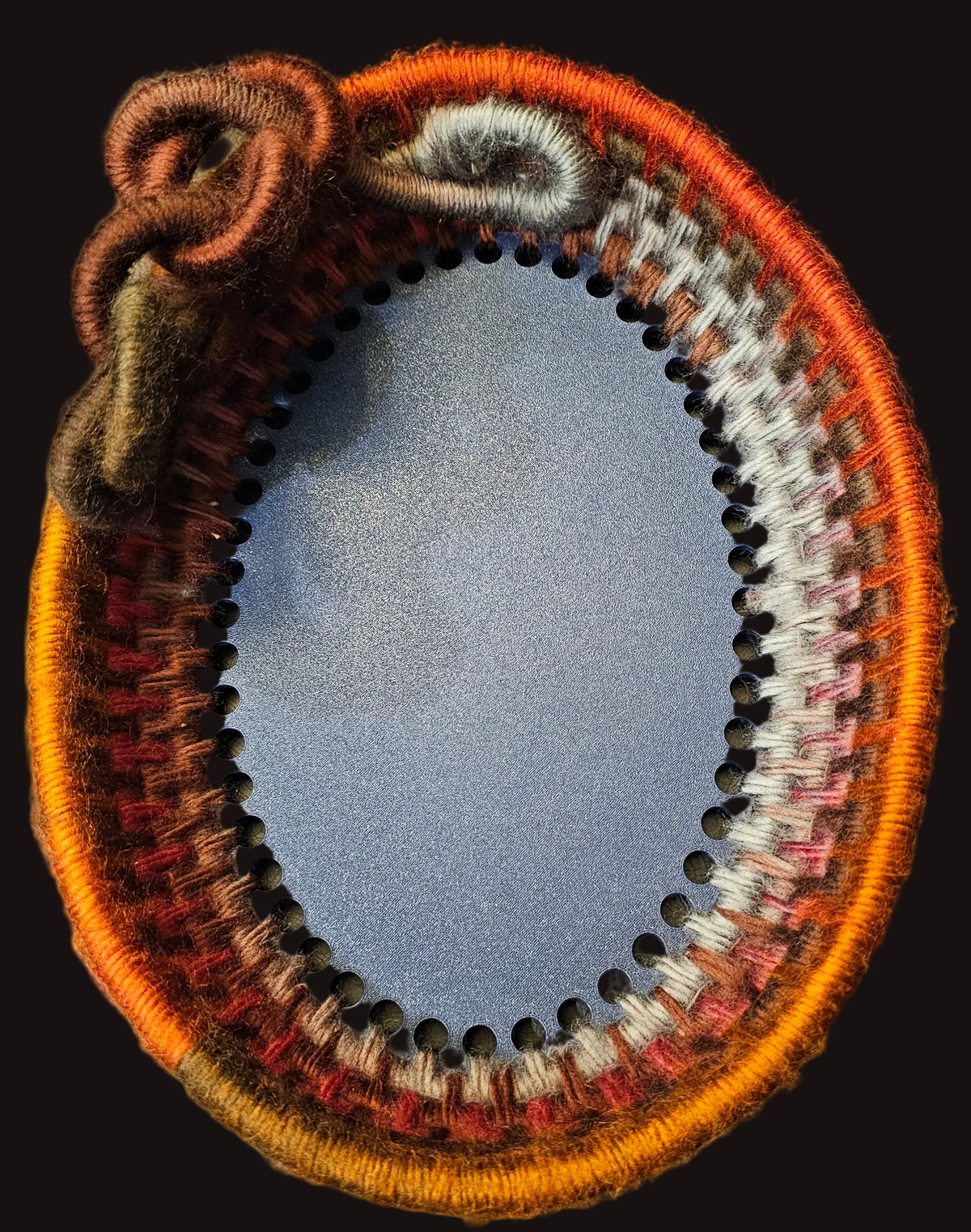 An oval handwoven basket made from variegated fuzzy yarn in deep, warm oranges, browns, reds, and gray; featuring an intricate knotted handle on the upper left.