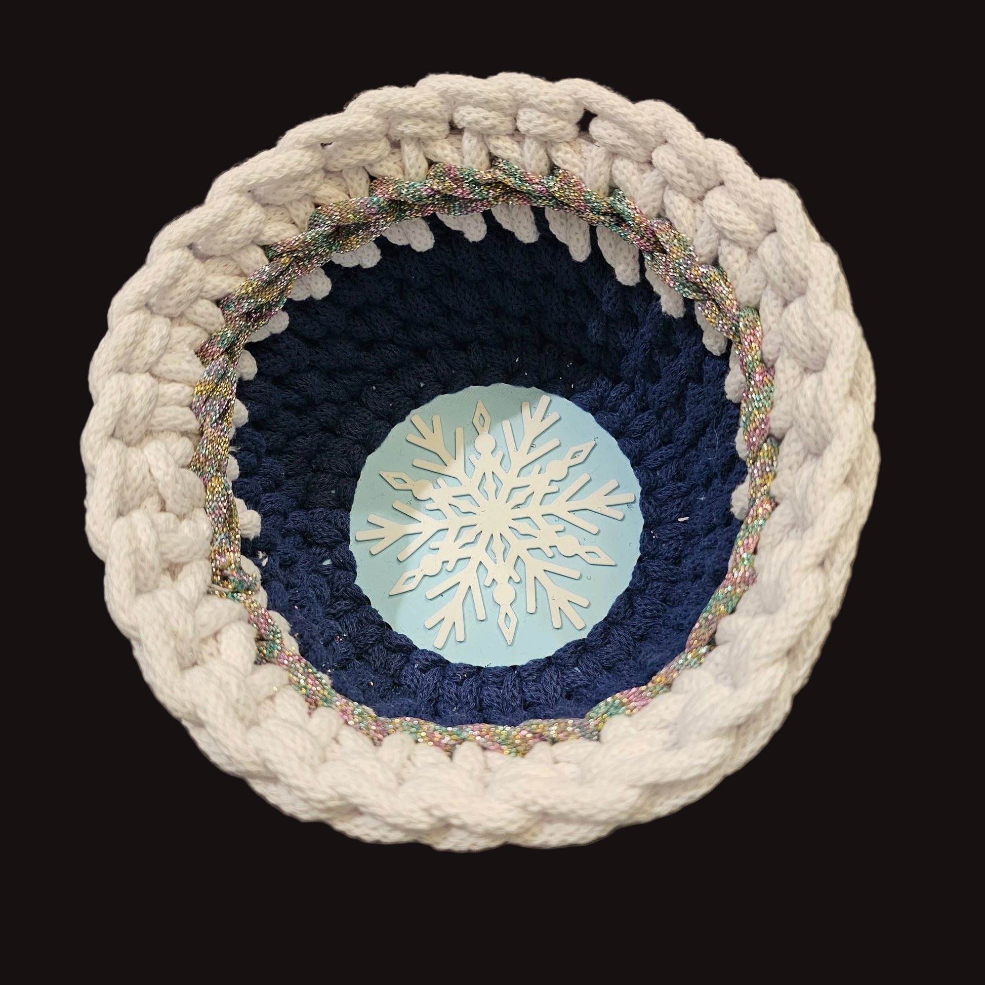 A round handmade crocheted basket with concentric rings of white chunky yarn, navy blue yarn, and a glittery multicolor border, featuring a light blue center with a white snowflake design.