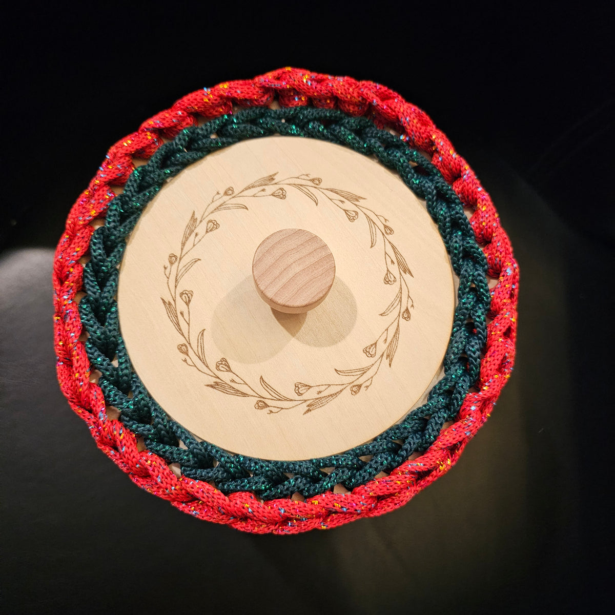 A rich crocheted lidded basket in candy apple red with a dark green tree motif on the front. Red and green metallic accents. The wooden lid has a natural wood grain finish and a central knob, decorated with an engraved floral wreath. 6x6x6 inches overall (4 inch center opening).
