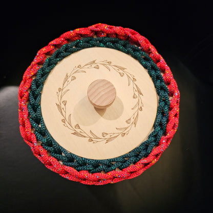 A rich crocheted lidded basket in candy apple red with a dark green tree motif on the front. Red and green metallic accents. The wooden lid has a natural wood grain finish and a central knob, decorated with an engraved floral wreath. 6x6x6 inches overall (4 inch center opening).