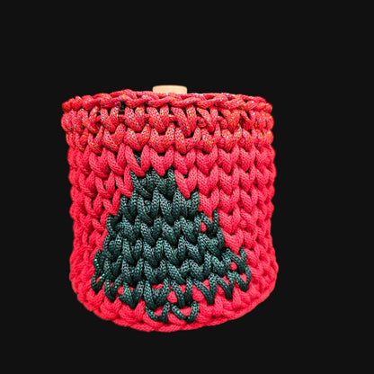 A rich crocheted lidded basket in candy apple red with a dark green tree motif on the front. Red and green metallic accents. The wooden lid has a natural wood grain finish and a central knob, decorated with an engraved floral wreath. 6x6x6 inches overall (4 inch center opening).