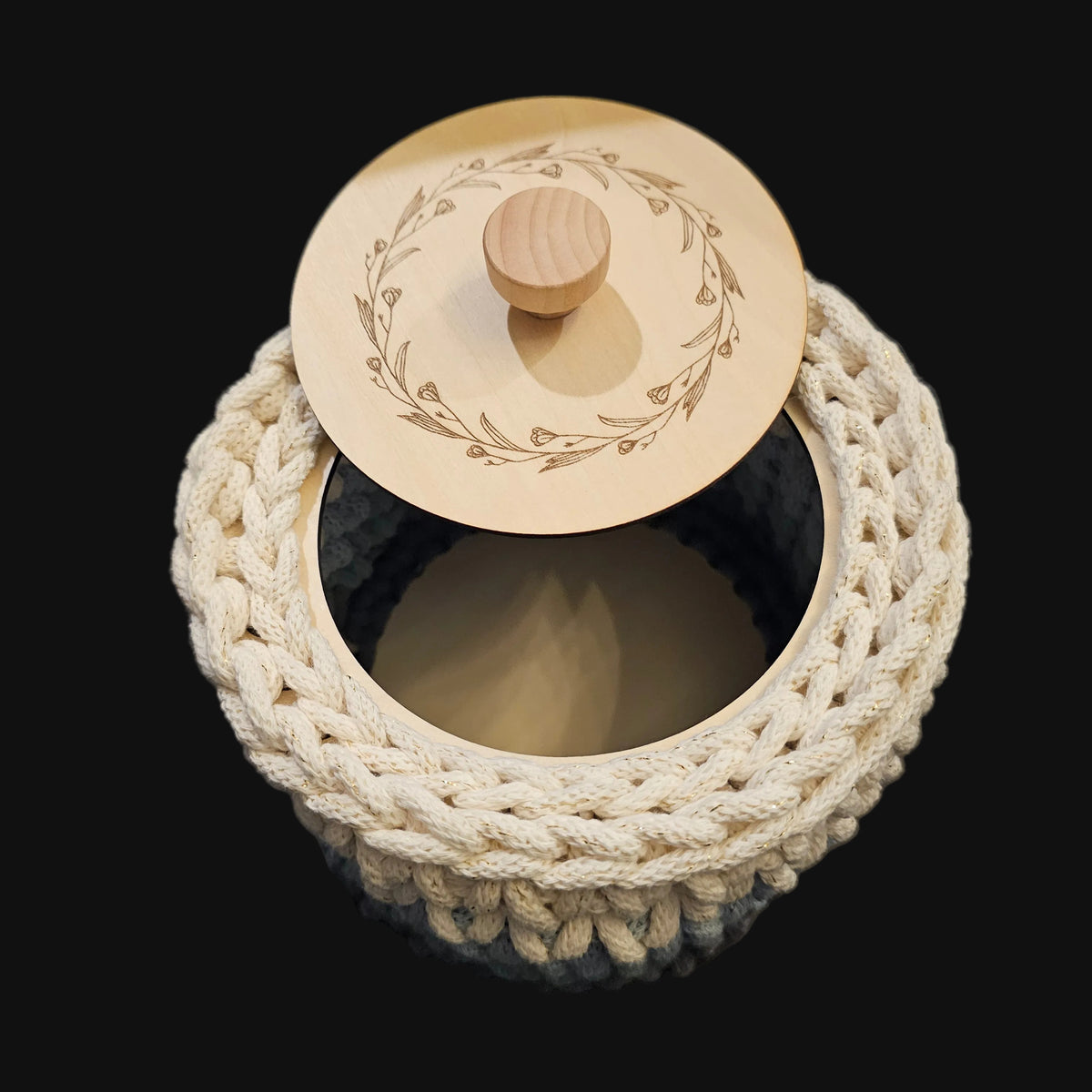 A round crocheted basket in cream (with flecks of gold thread), light blue, and gray with a matching round wooden lid featuring an engraved floral wreath design and a small wooden knob. 6x6x5 inches overall; basket opening is 4 inches in diameter.