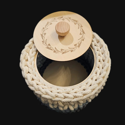 A round crocheted basket in cream (with flecks of gold thread), light blue, and gray with a matching round wooden lid featuring an engraved floral wreath design and a small wooden knob. 6x6x5 inches overall; basket opening is 4 inches in diameter.