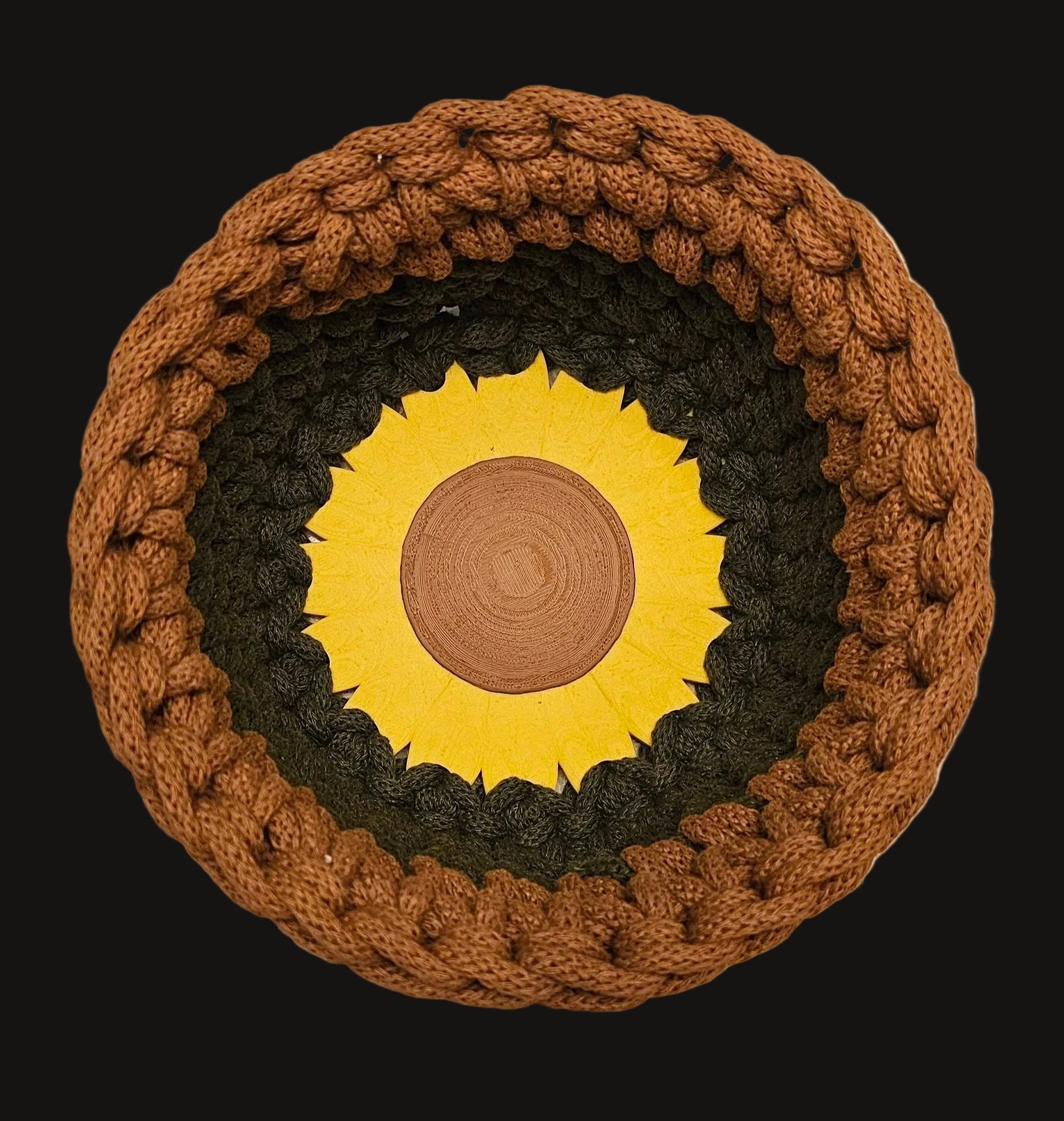 A crocheted basket with a bright yellow sunflower at its center. This basket is a two-tone design, featuring a warm brown top section and an olive-green bottom section. 7x7x4 inches overall