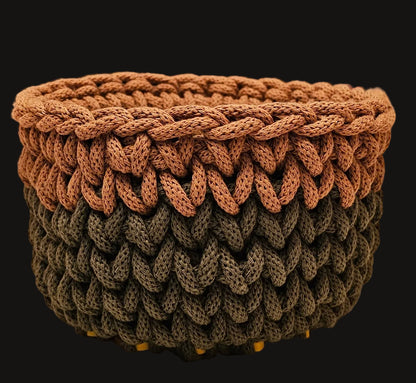 A handmade braided basket with a two-tone design, featuring a warm brown top section and an olive green bottom section, resting on a light speckled countertop.