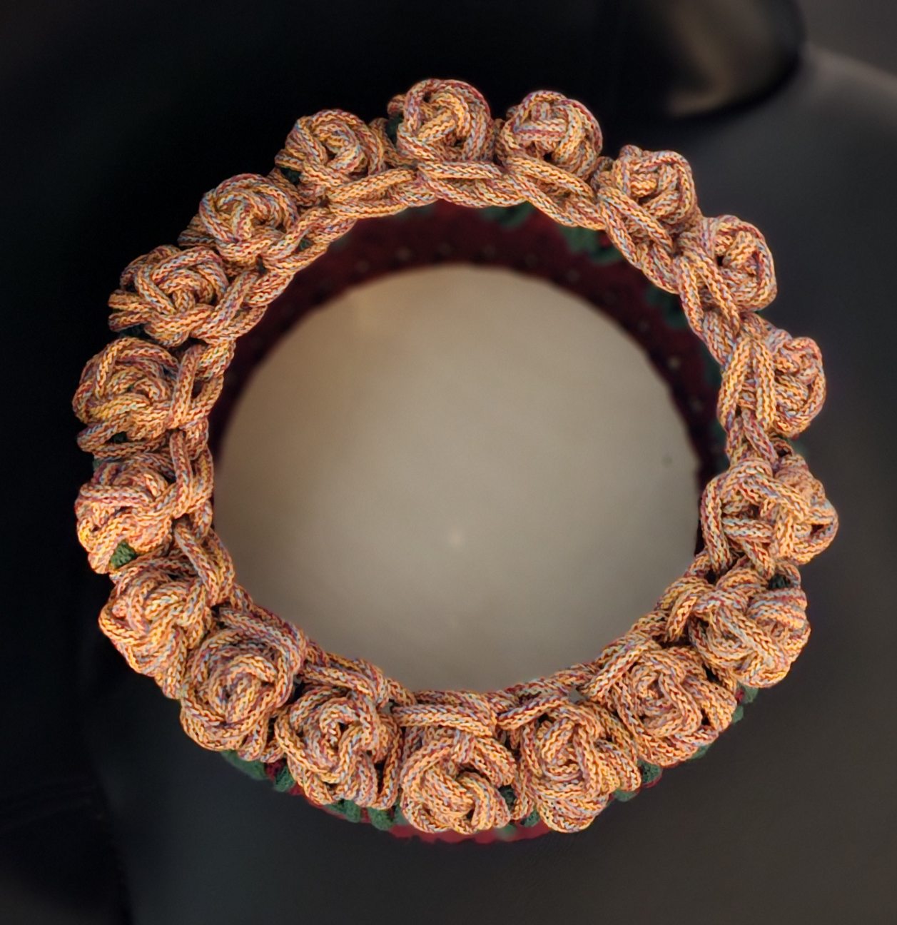 A handmade crocheted basket with a marled orange, blue and red flower like rim, with dark green leaves, and a deep wine base. All secured to a round wooden base. 9x9x5 inches overall.
