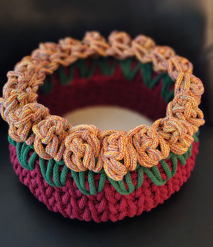 A handmade crocheted basket with a marled orange, blue and red flower like rim, with dark green leaves, and a deep wine base. All secured to a round wooden base. 9x9x5 inches overall.