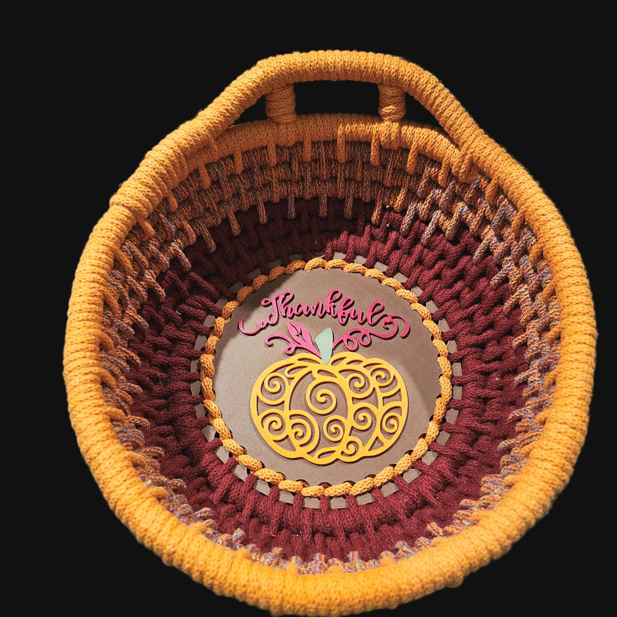 A coiled basket with a top handle, featuring a striped pattern in orange, terracotta, and burgundy. The center is  a yellow pumpkin design and the word 'Thankful' in burgundy script. 9x9x4 inches overall.
