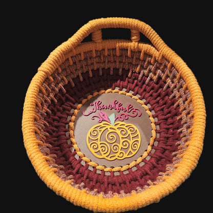 A coiled basket with a top handle, featuring a striped pattern in orange, terracotta, and burgundy. The center is  a yellow pumpkin design and the word 'Thankful' in burgundy script. 9x9x4 inches overall.